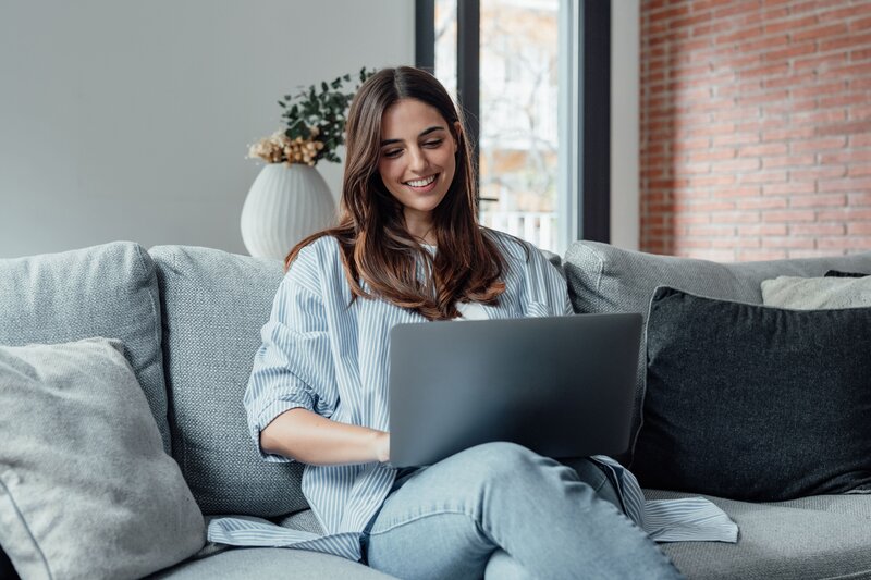 Frau sitzt entspannt auf dem Sofa und arbeitet lächelnd mit einem Laptop.