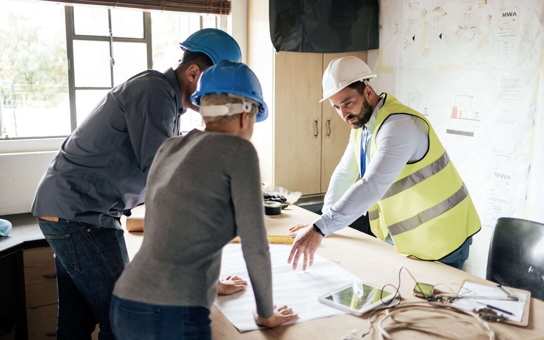 Mehrere Personen mit Schutzhelmen besprechen Baupläne an einem Tisch auf einer Baustelle.